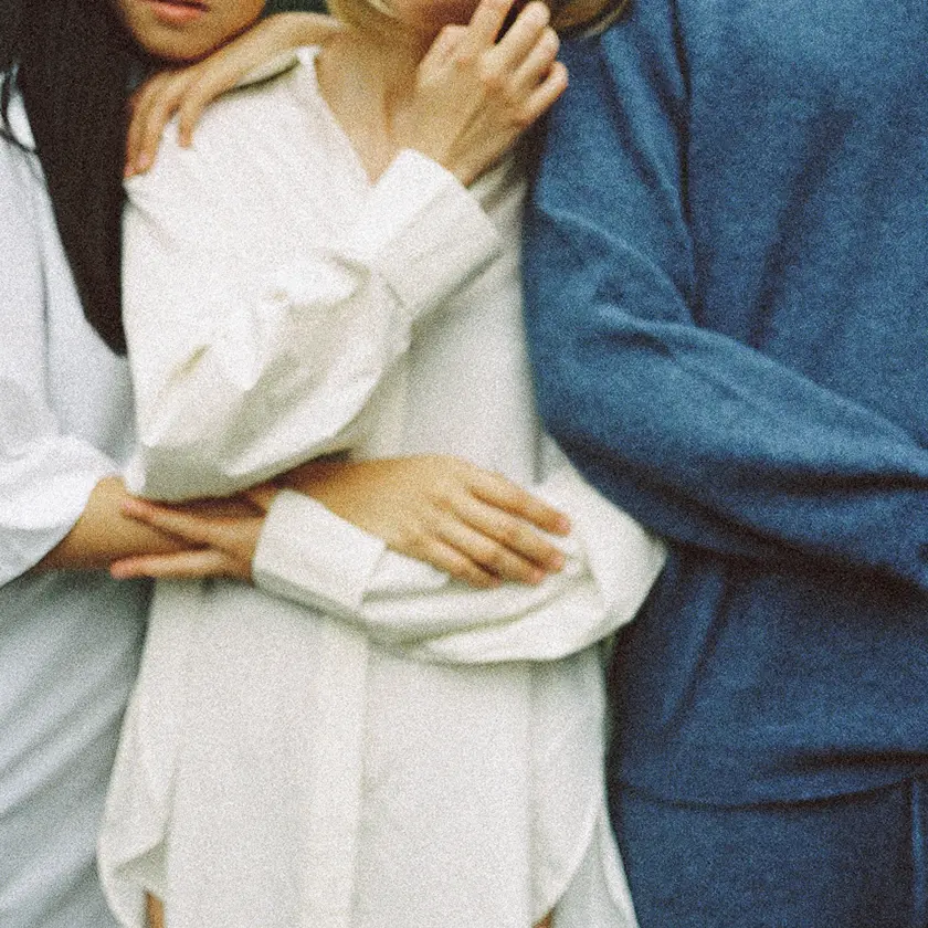 Three people are lying on the grass, enjoying a sunny day. The background includes trees and a clear sky.
